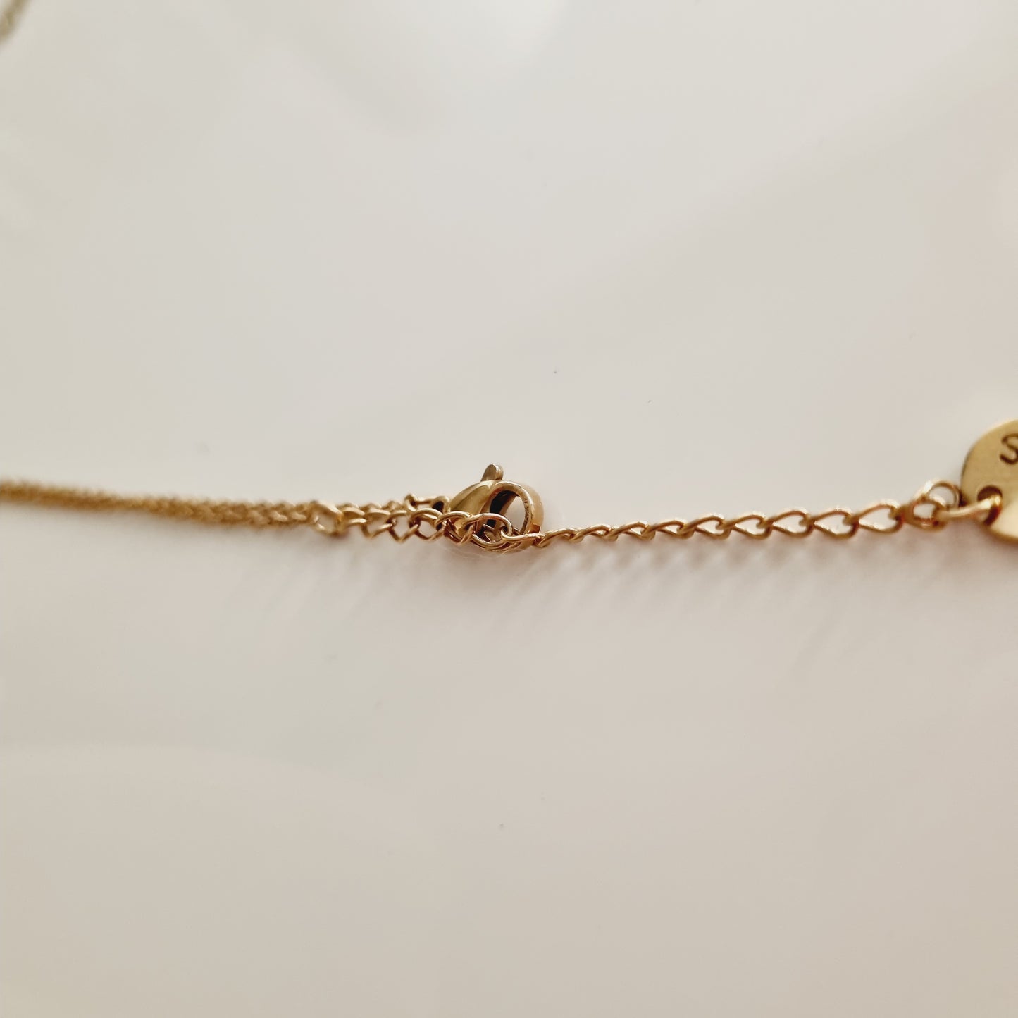 Zoom sur le fermoir et l’extrémité d’une chaîne en acier doré à maillons fins, photographiée sur fond neutre. Un détail délicat qui évoque la qualité et la finition soignée, parfait comme cadeau discret mais élégant pour un anniversaire, Noël ou un remerciement raffiné. La personne qui la reçoit ressent un sentiment de douceur, de raffinement et d’attention portée aux détails.