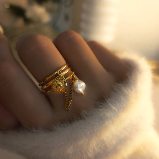 Bague multi-anneaux dorée avec pendentif soleil, perle et chaîne fine portée à la main