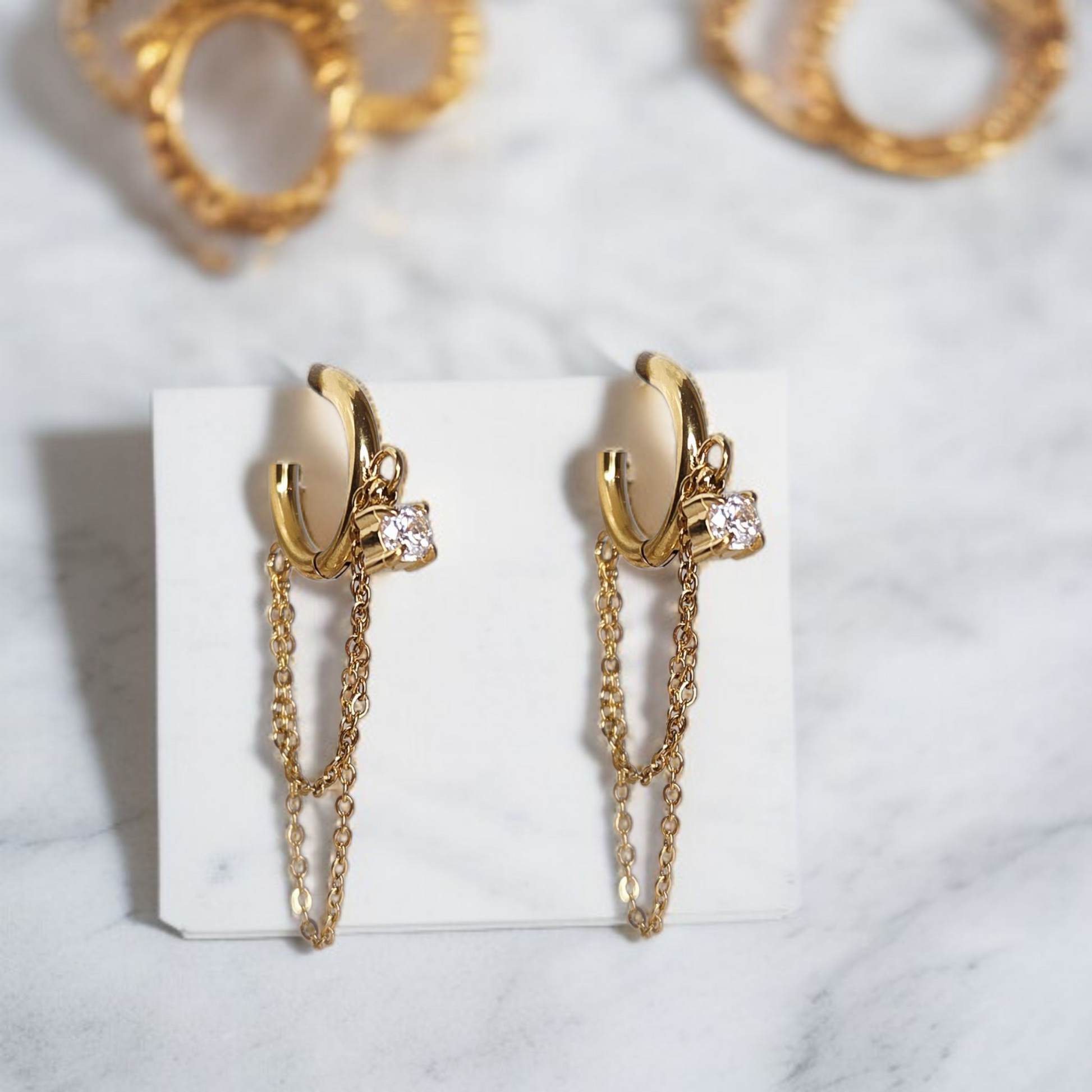 Boucles d'oreilles dorées sur fond clair présentoir blanc. Avec 2 chaînes pendantes, effet très classe.