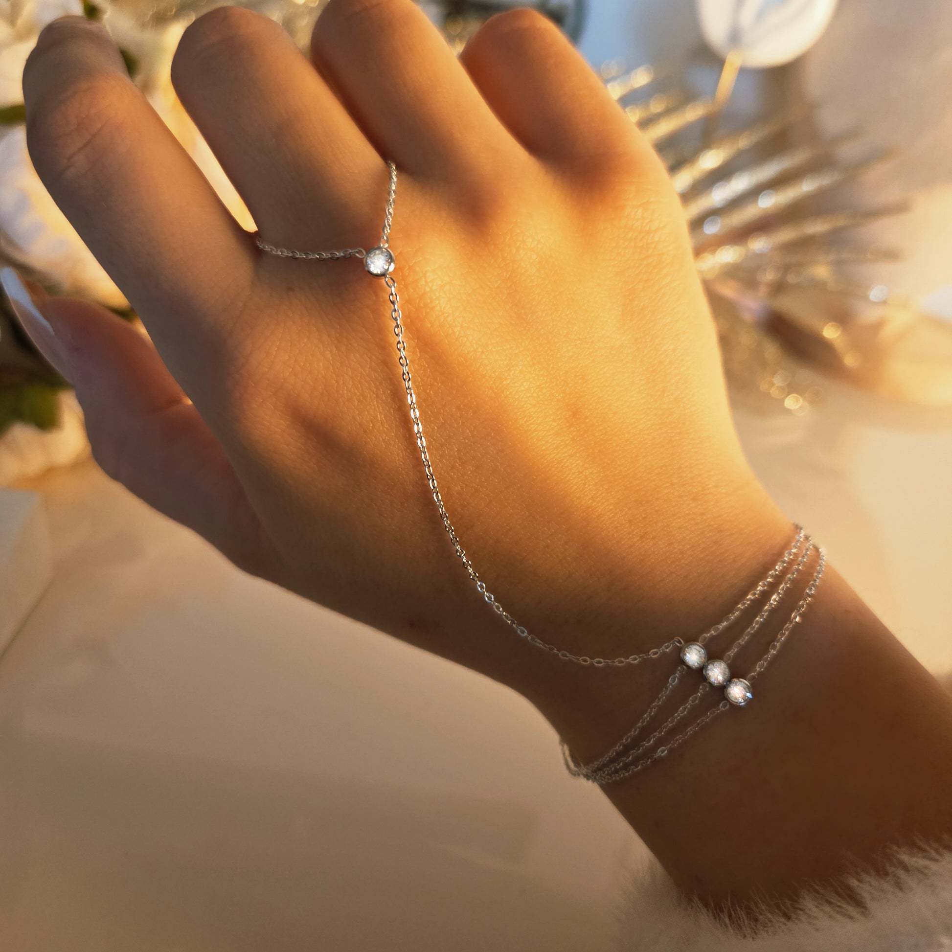 Le bijou de main est porté sur la main, révélant l’harmonie entre la chaîne reliant le doigt et le bracelet au poignet. La finesse du design souligne élégamment la main tout en mettant en valeur les détails lumineux.