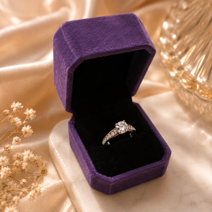 Bague solitaire argentée avec pierre centrale ronde et pierres blanches latérales présentée dans un écrin violet