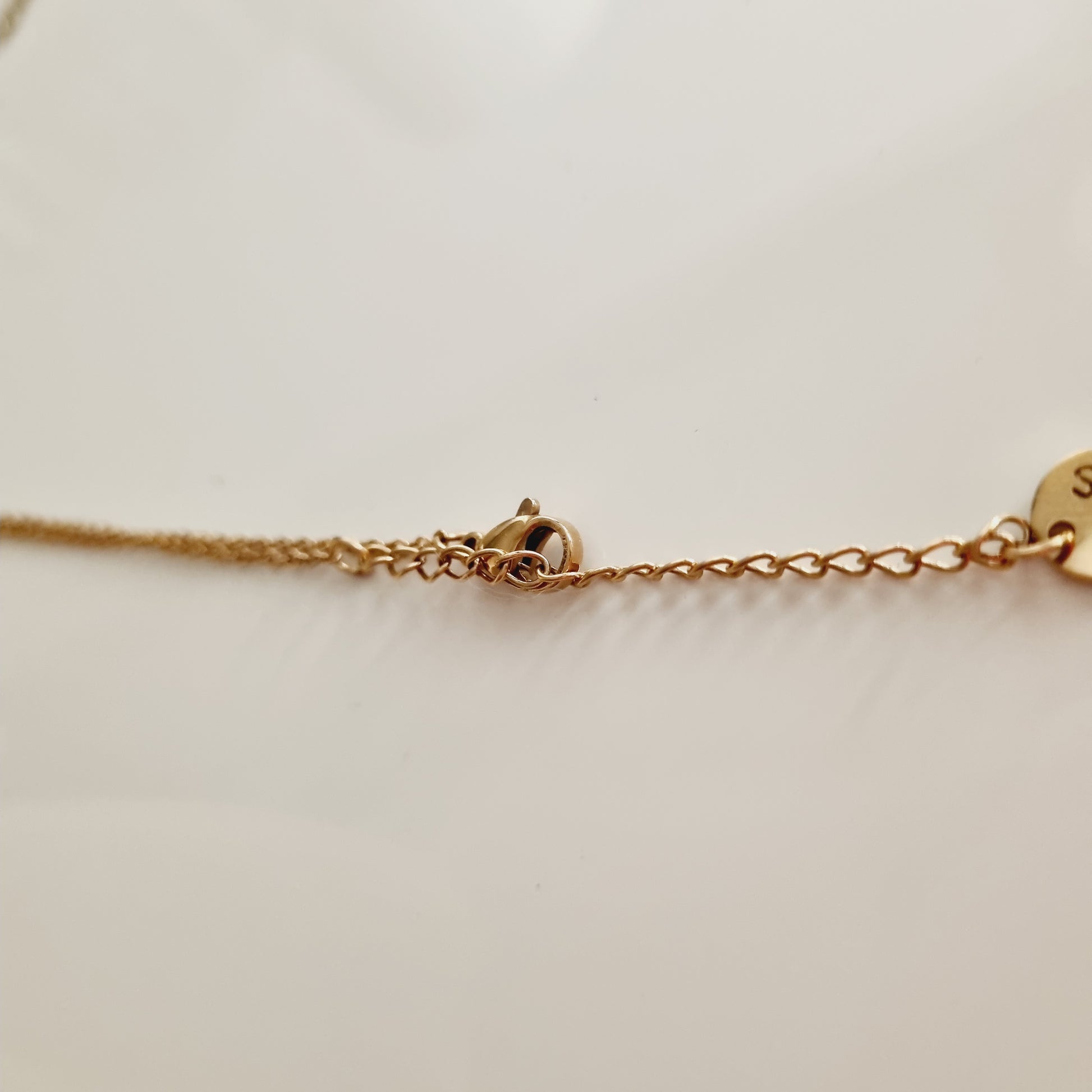 Zoom sur le fermoir et l’extrémité d’une chaîne en acier doré à maillons fins, photographiée sur fond neutre. Un détail délicat qui évoque la qualité et la finition soignée, parfait comme cadeau discret mais élégant pour un anniversaire, Noël ou un remerciement raffiné. La personne qui la reçoit ressent un sentiment de douceur, de raffinement et d’attention portée aux détails.